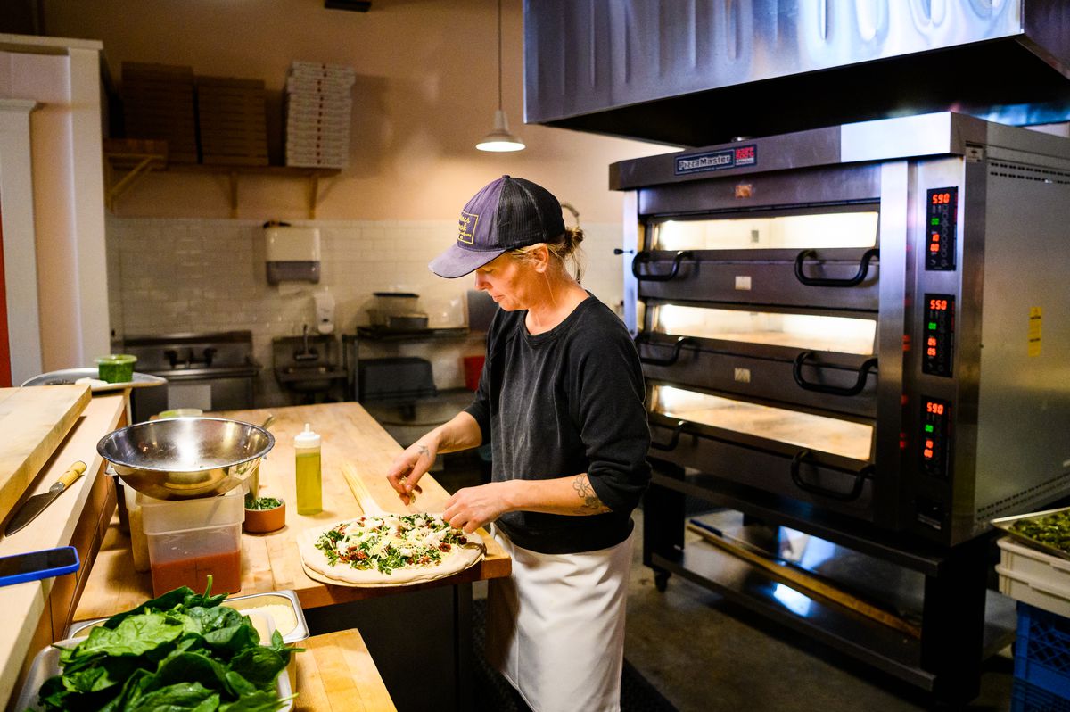A woman in a baseball cap and apron puts toppings on an uncooked pie, with a deck oven behind her.