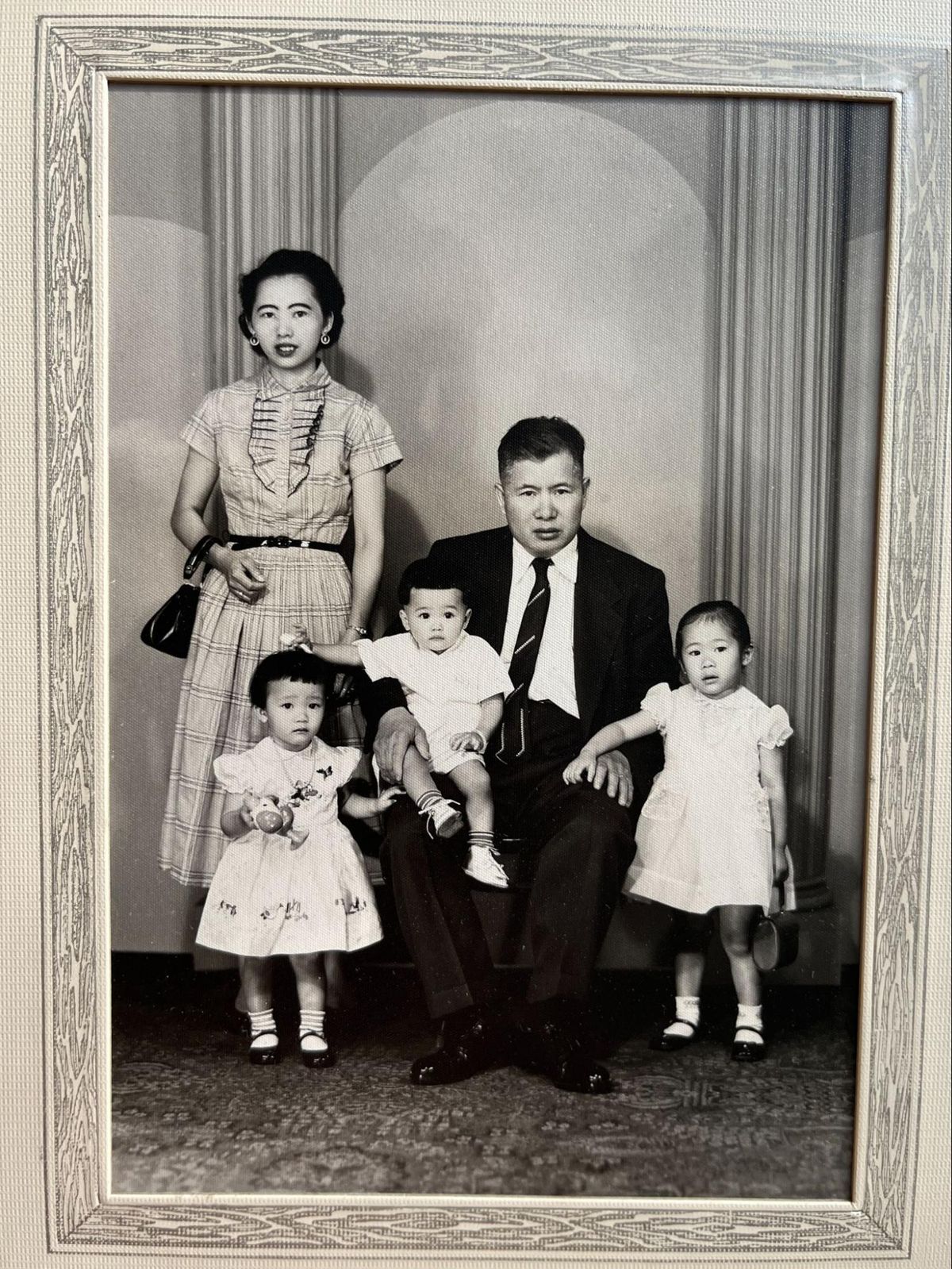 A woman in a plaid dress and holding a black bag standing to the left, a man in a black jacket, dark tie, white shirt sitting holding a baby, two small children in dresses standing in front.