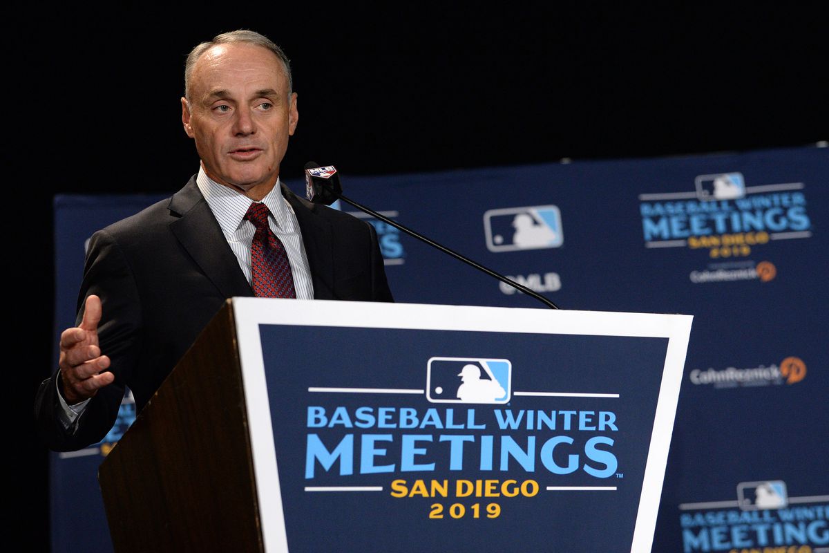 MLB: Rob Manfred speaks from a Winter Meetings podium, probably explaining how the most baseball players get hospitalized with COVID-19, the bigger bonus he’ll get
