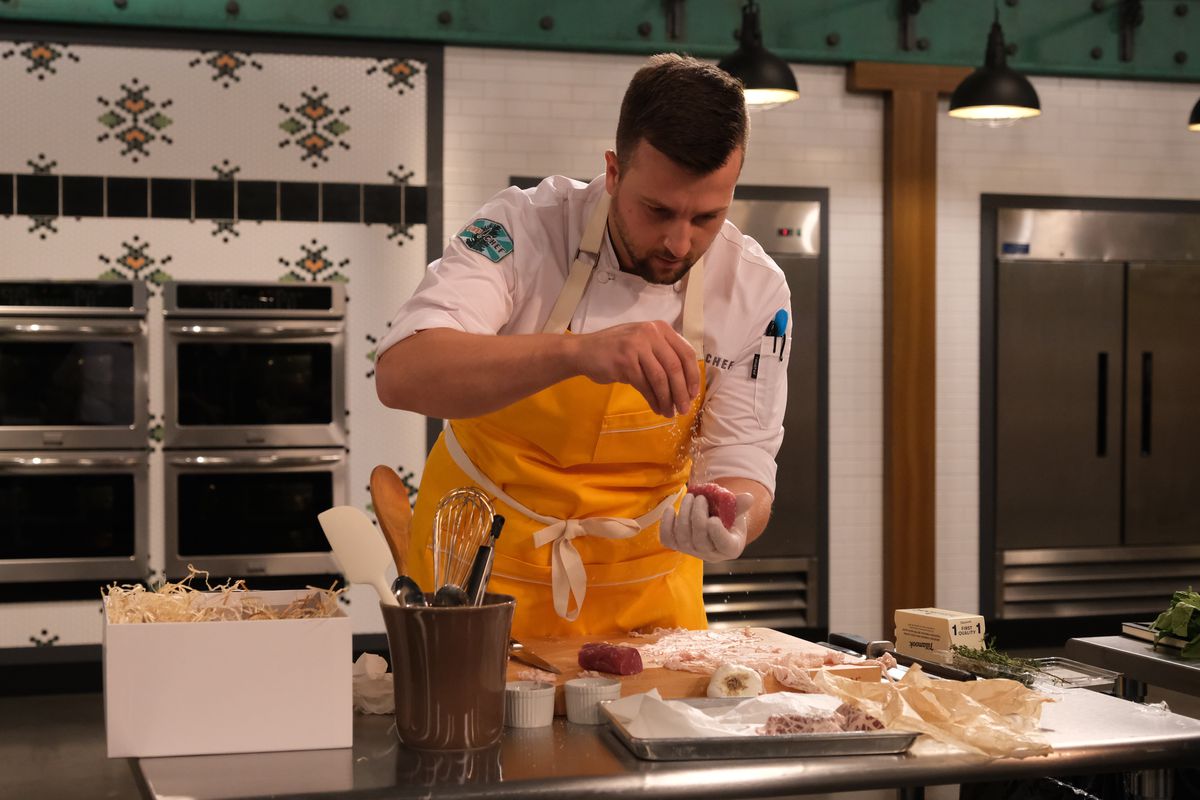 Gabriel Pascuzzi seasons a plate of food wearing a yellow apron in a white kitchen. He’s leaning over the food in chef’s whites.