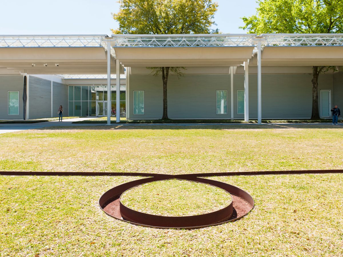 The exterior of the Menil Collection in Texas. The building is rectangular with steel support beams. In the foreground is a lawn with a round steel cut out design in the center.