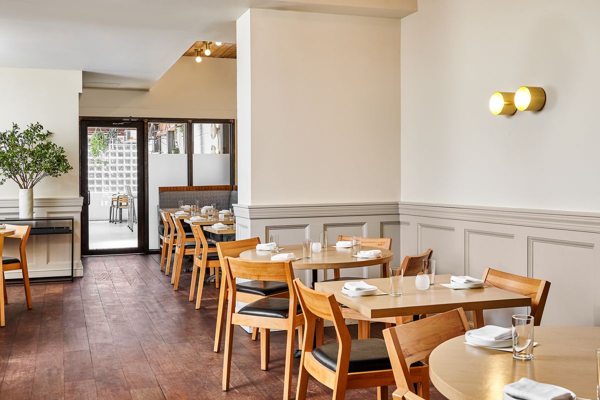The indoor dining room of a restaurant is outfitted with light wood furniture, white walls, and tables set for service.