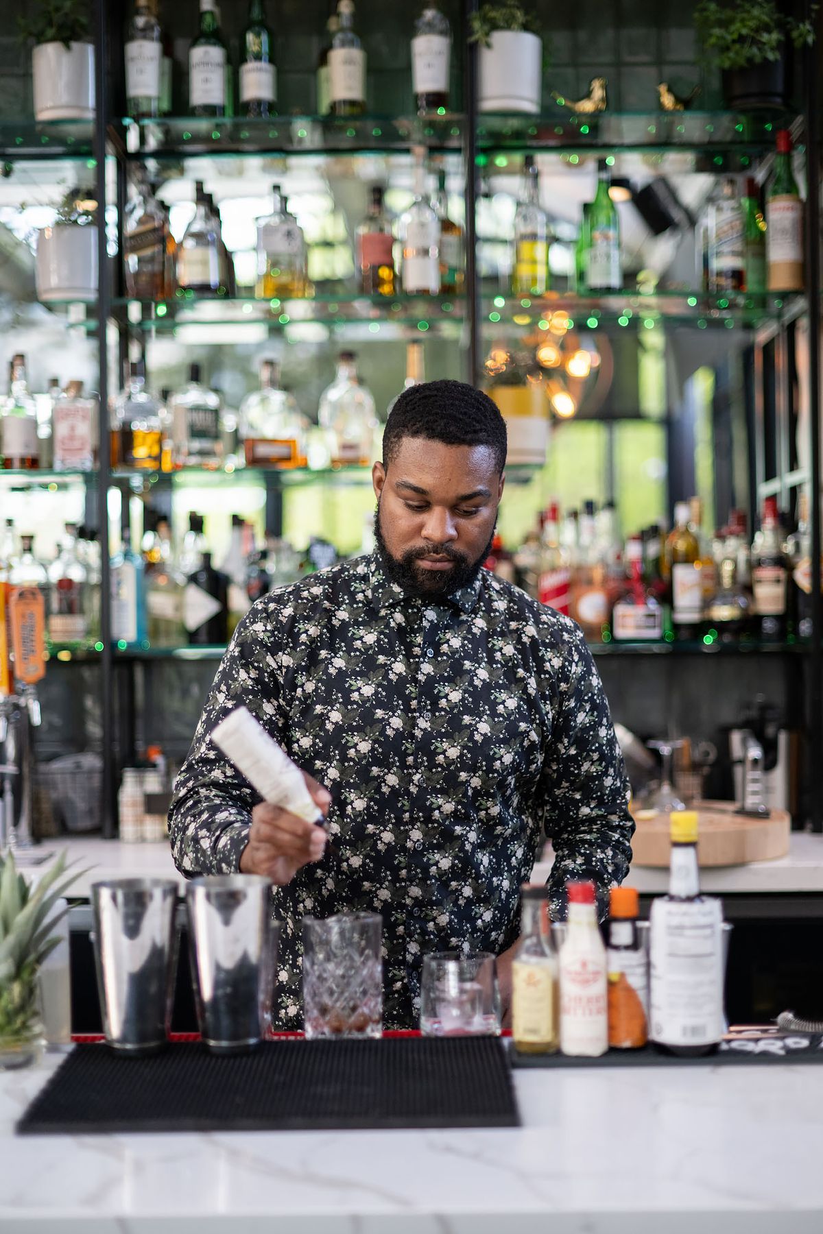 A bartender in a floral print shirt pours dashes of bitters into a cocktail.