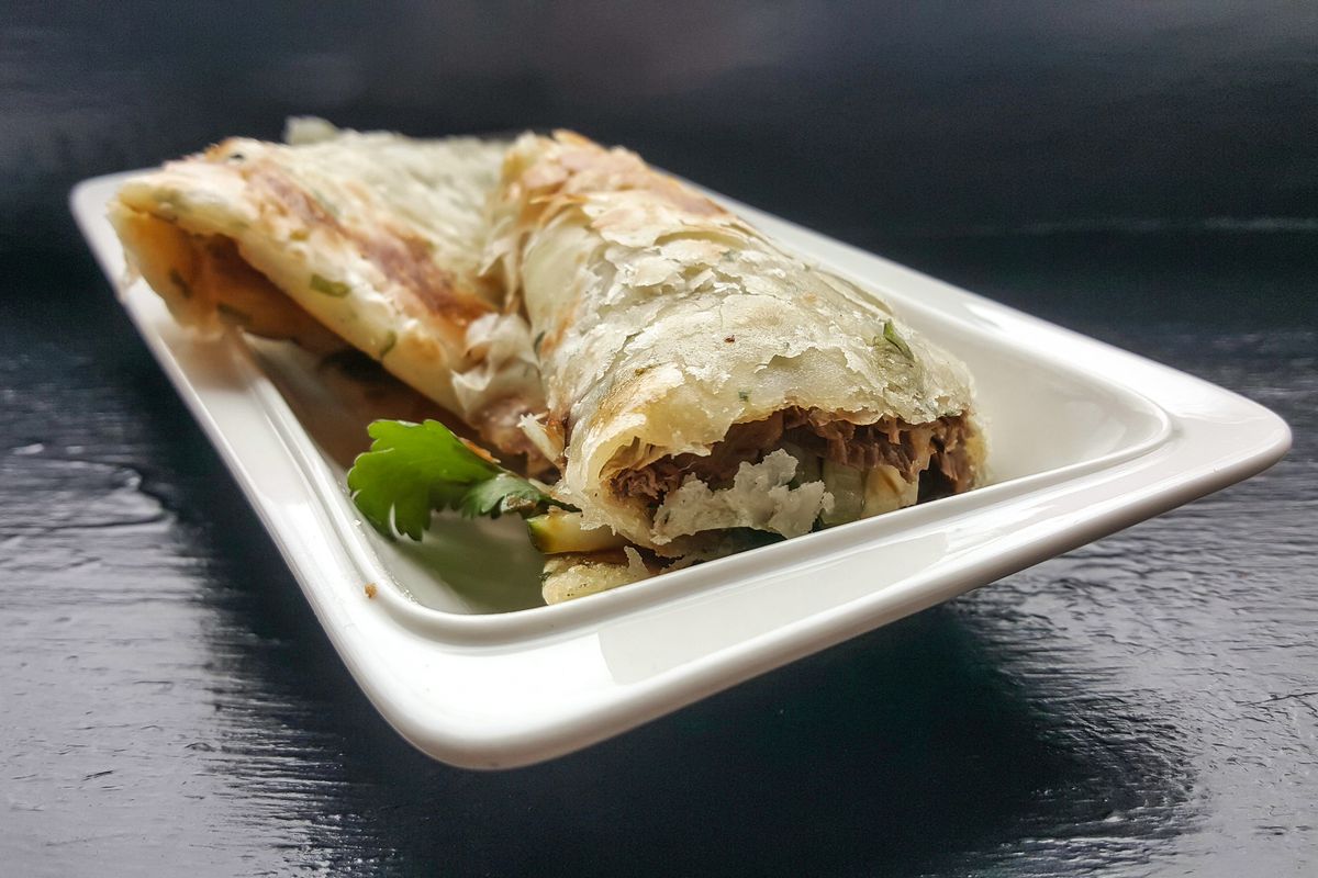 Beef is wrapped in a flaky scallion pancake, sitting on a white plate on a black tabletop