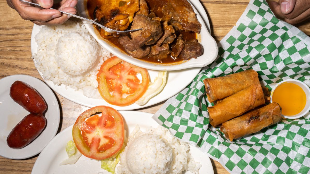 A hand spoons red adobo from a white dish alongside a collection of other dishes including white rice, lumpia, and tomatoes. It all sits on a green and white checked tablecloth.