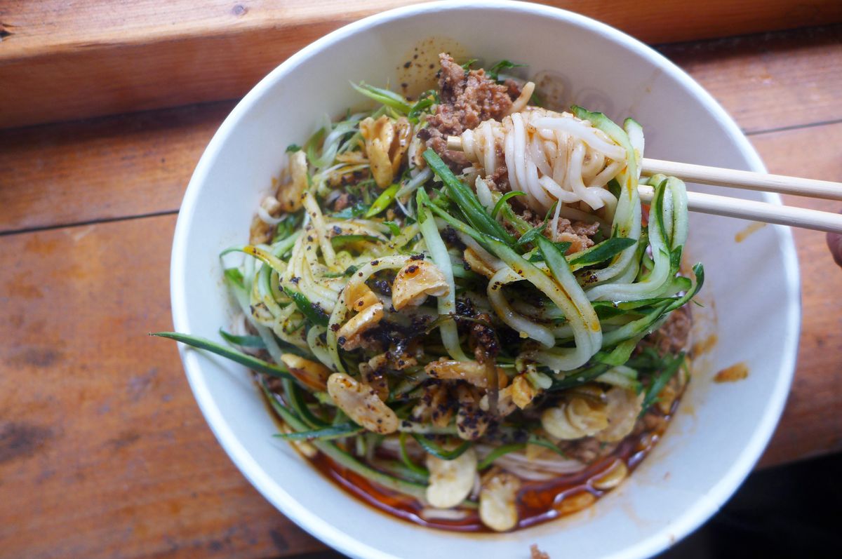 A pair of chopstick pull a hank of noodles out of a paper receptacle against a backdrop of slivered cucumbers, ground pork, and crush chiles.