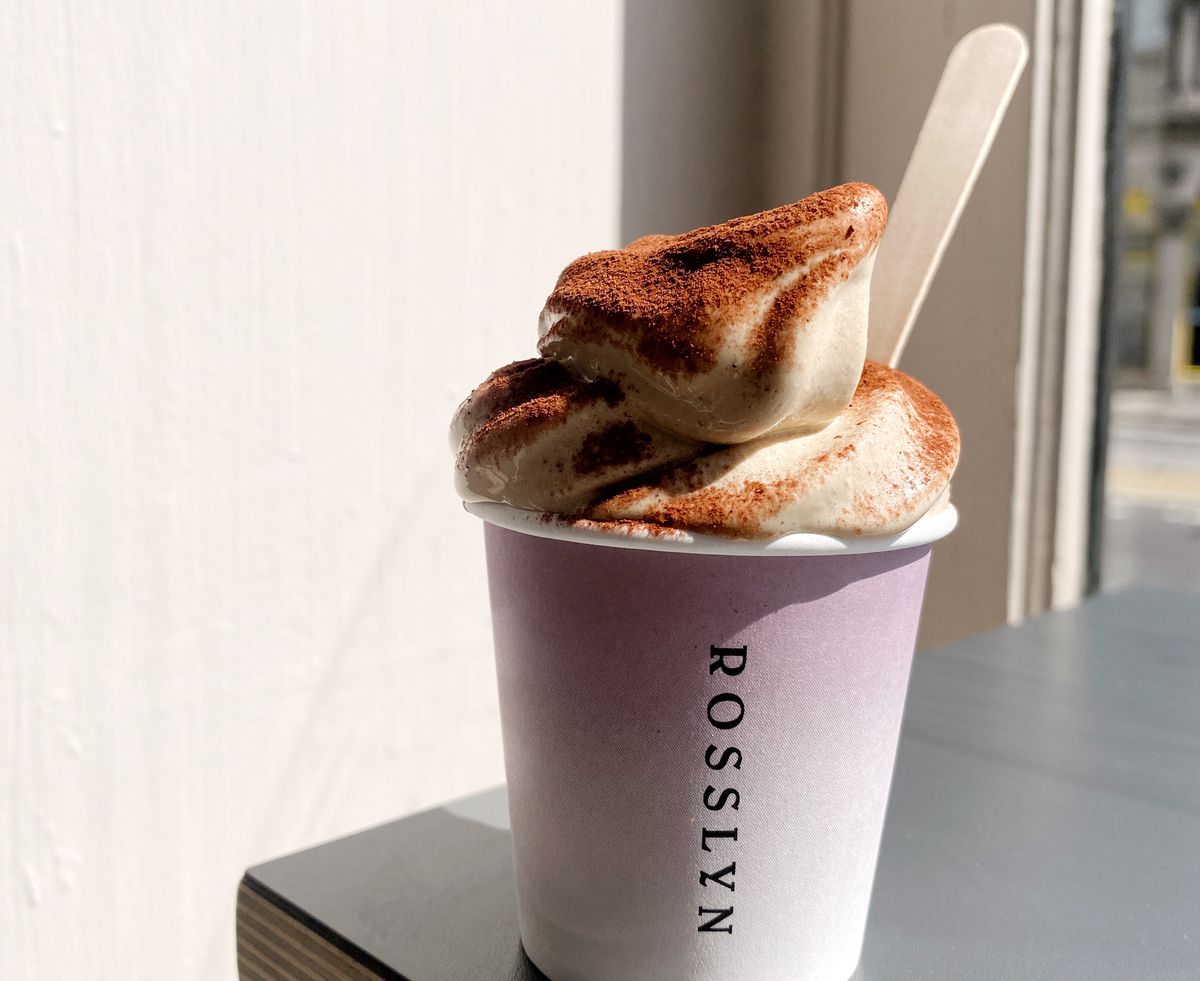 Espresso soft serve served in a purple takeaway cup with a small wooden ice cream spoon, with chocolate powder on top