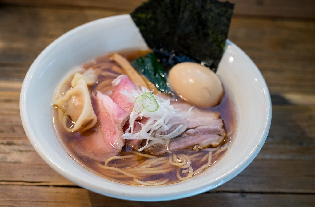 A bowl of ramen with relatively thin noodles, slices of pork stacked above the surface, a wonton, bamboo shoots, a slice of seaweed sticking up, and a boiled egg.