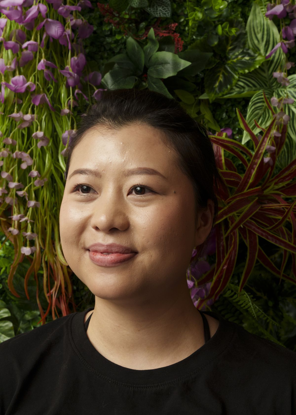 Aunyamanee Ritneatikun, wearing a black shirt with her hair pulled back, smiles and looks somewhere past the camera as she stands in front of green foliage and purple flowers. 
