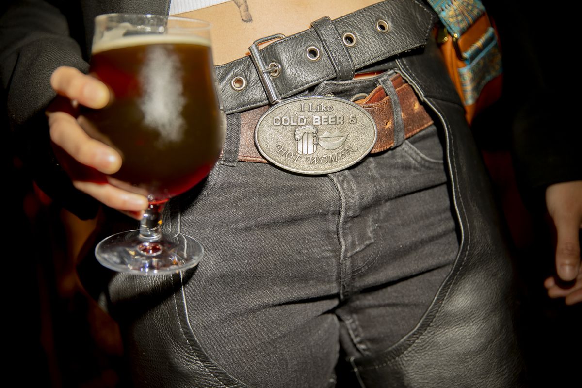 Shot of waist and thighs. A buckle depicts lips and a beer stein along with text. Part of a tattoo peeks out under shirt hem.