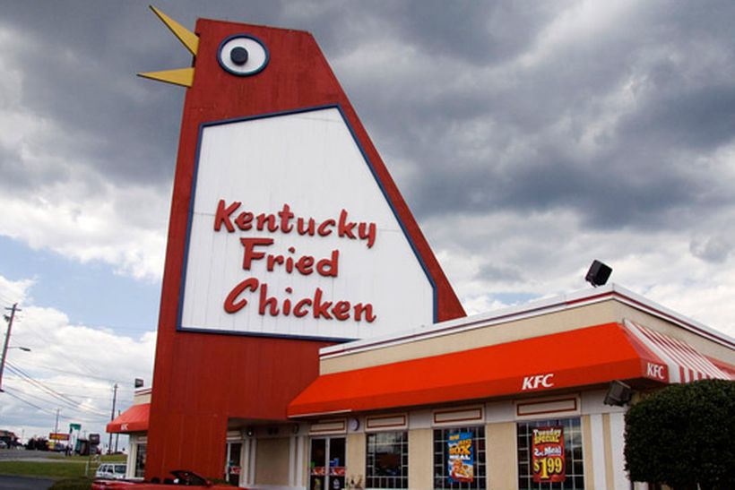 A building with a giant red chicken on the roof. There is a sign on the chicken that reads: Kentucky Fried Chicken. There is a red awning on the building.