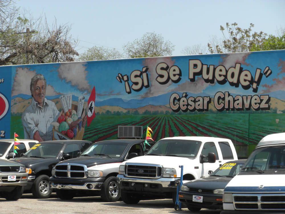 A mural of Cesar Chavez that says "Si Se Puede" with a car lot in front