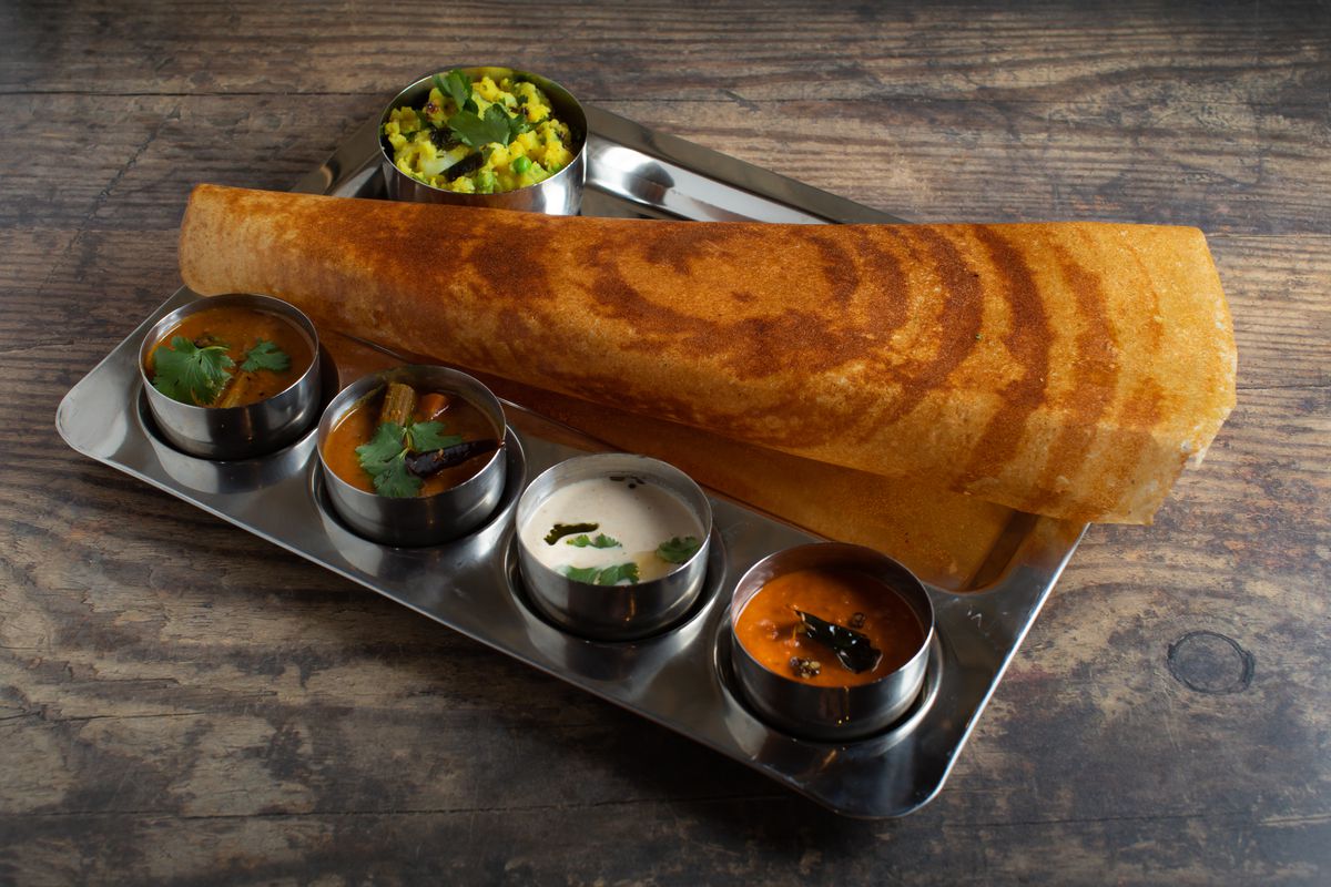 A long, thin dosa sits on a silver tray on a wooden surface. The tray also holds five small silver bowls of chutneys and sauces.