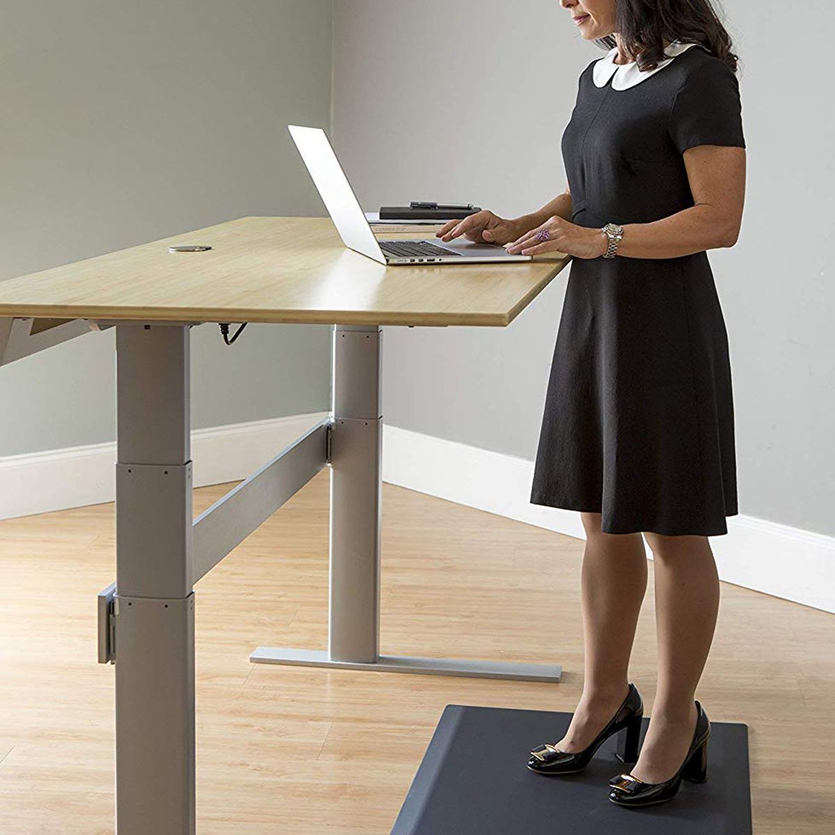A woman wearing a black desk stands on a gray mat while working at a birch standing desk. 