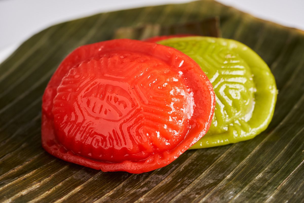 Mai multe angku kuih roșii și verzi imprimate cu modele de coajă de țestoasă, aranjate pe o frunză de banană.