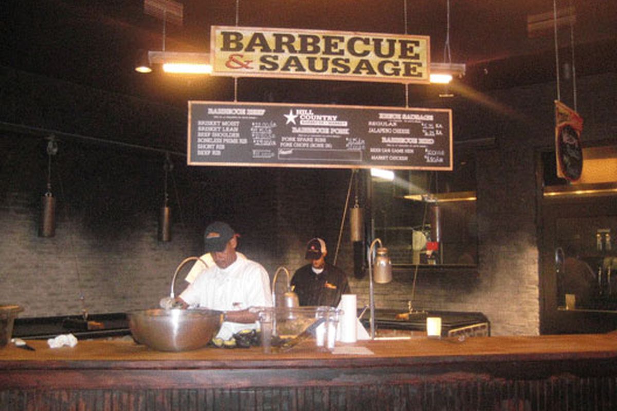 Two men stand behind a counter with a big sign up above advertising barbecue and sausage.