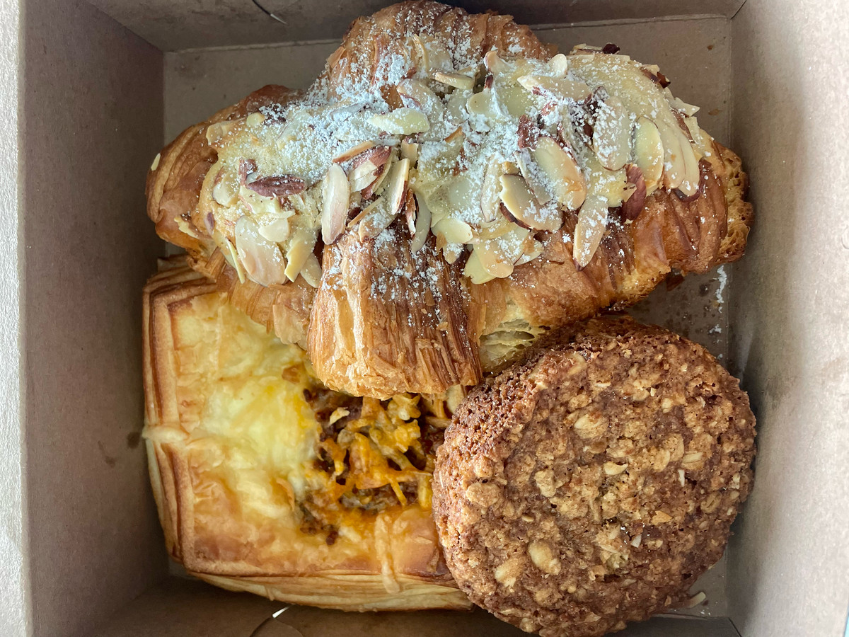 A pastry box holds an almond croissant, an oatmeal cookie, and a pastry with egg and chorizo.