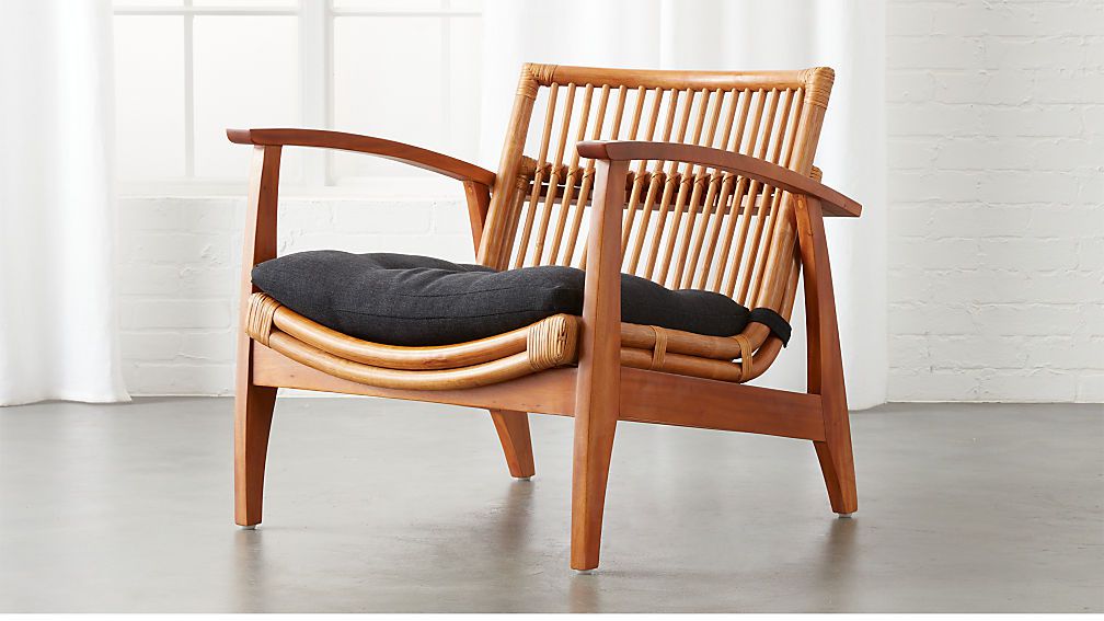 A rattan arm chair with an open-slated back, high arms, a curved seat, and black cushion. 
