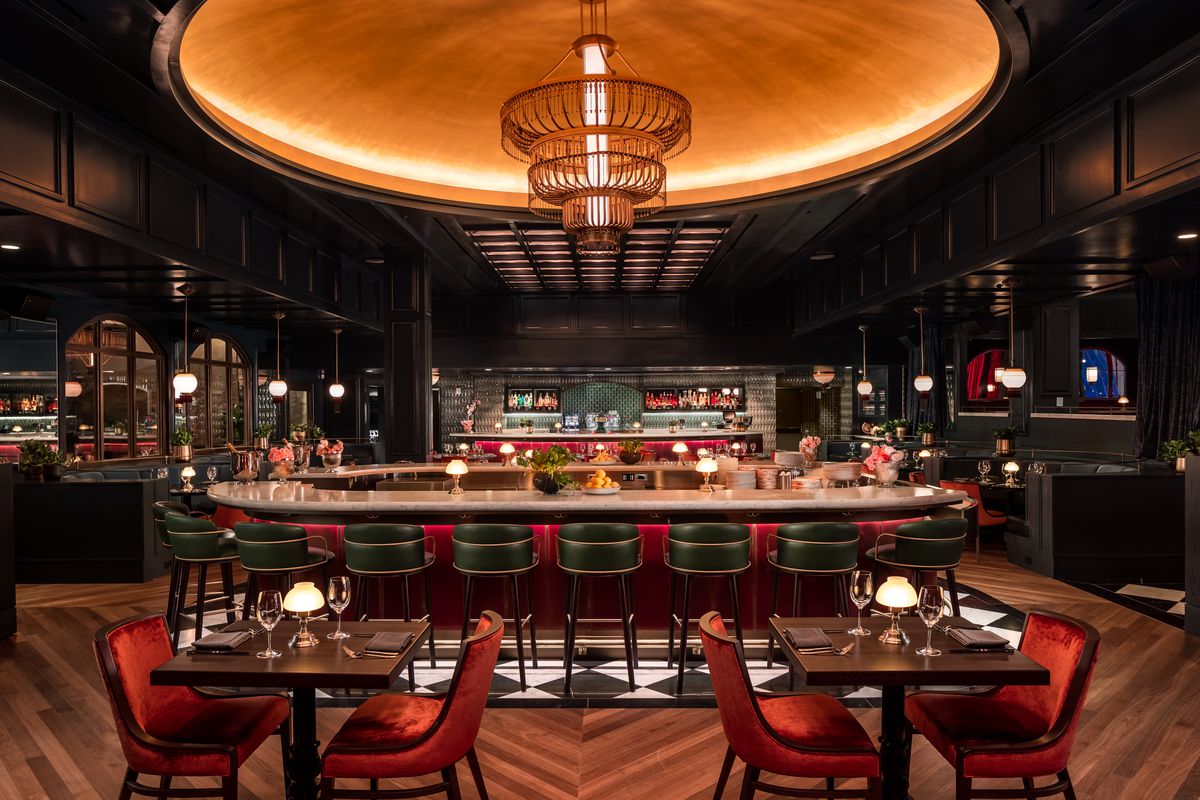 A wood paneled room with wood floors where the central focus point is the bar surrounded by black leather bar chairs and a massive chandelier overhead. 