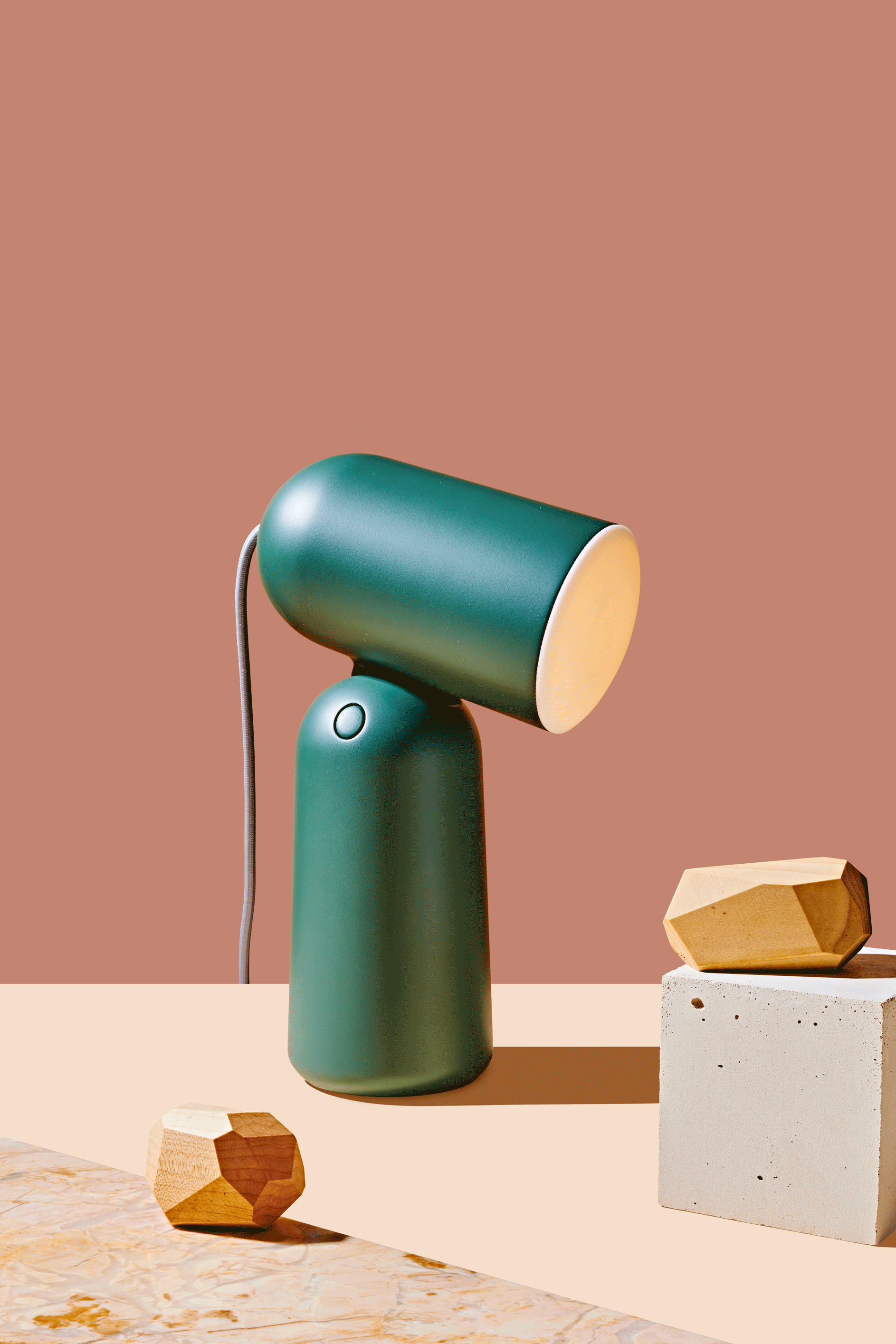 A dark green Buddy Table Light which is part of the 2019 Curbed Holiday Gift Guide. The top part of the light is moving up and down. This is an animated gif. On the table around the light are various design objects. There is a mauve backdrop.