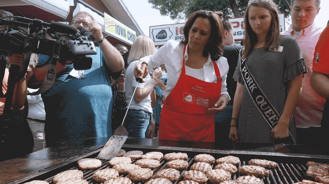 An animated GIF of Kamala Harris flipping pork chops on a large grill while surrounded by a crowd