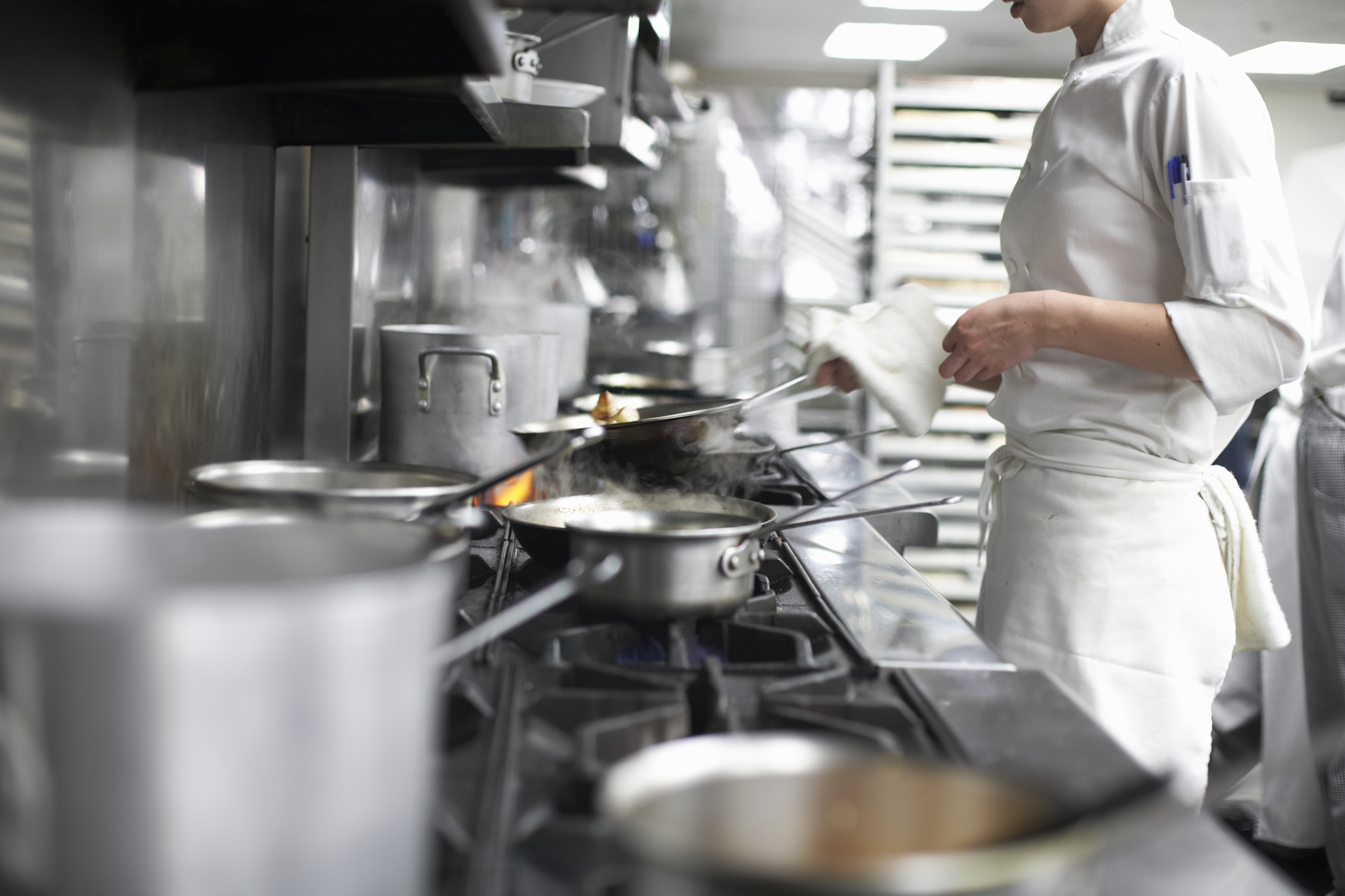 A female chef, dressed in white and visible from the chin down, stands over the stove in a restaurant kitchen