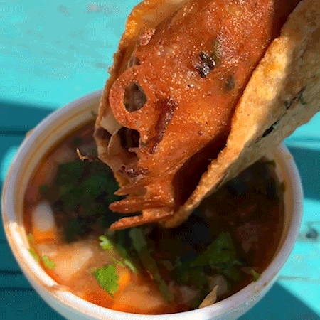 A hand dipping a quesataco with griddled cheese into a styrofoam cup of consomme on top of a teal wooden table