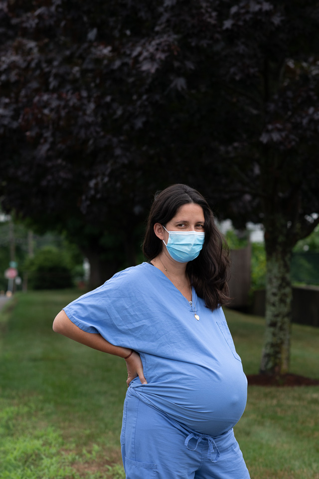 A pregnant woman wearing blue scrubs and a blue mask stands with a hand on her hip. She is framed by a line of trees out of focus behind her.