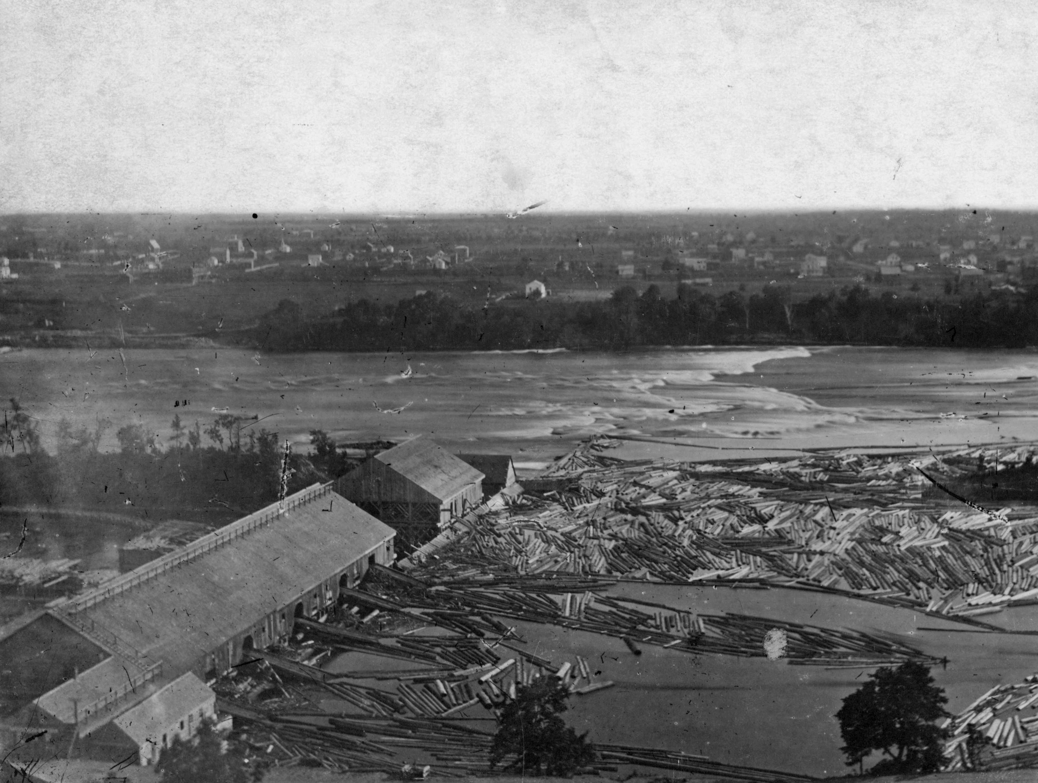 A sawmill on the Mississippi River across from downtown Minneapolis in the 1860s or 1870s processed logs that are floating in the water.