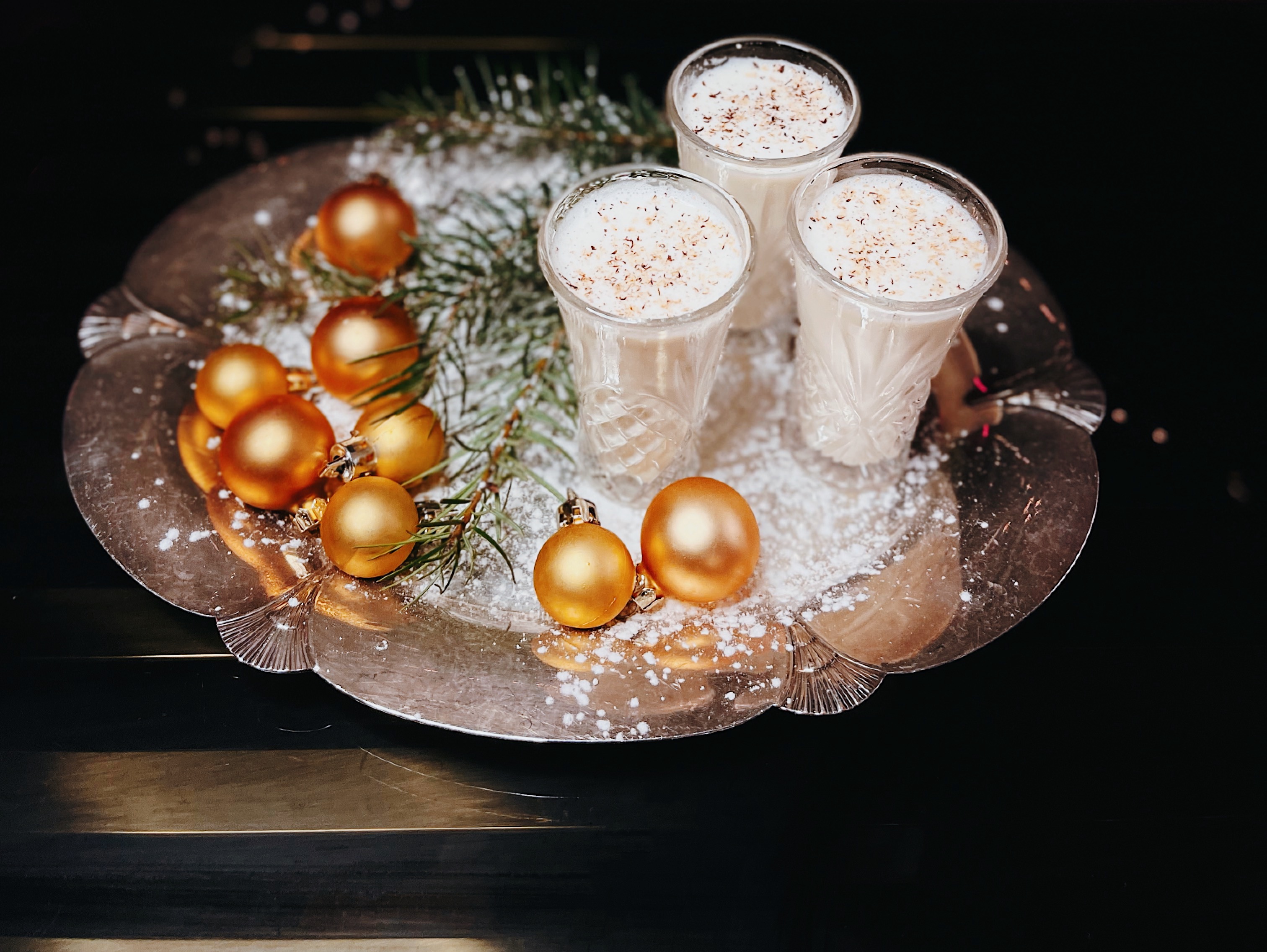 Three glasses of eggnog sit on a tray next to Christmas ornaments.
