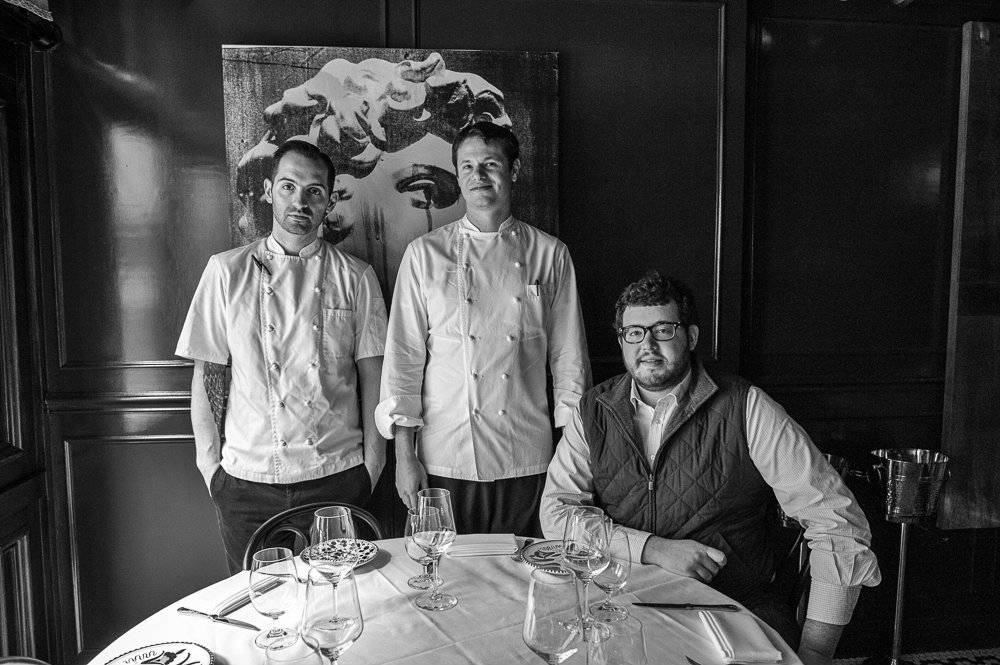 A black-and-white photo of Mario Carbone and Rich Torrisi, who are standing and wearing chef’s whites, and a sitting Jeff Zalaznick, who wears a vest.