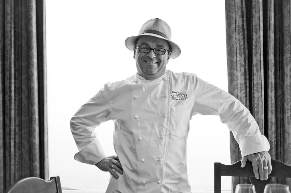 A black-and-white photo of a smiling man in a chef coat and a hat.