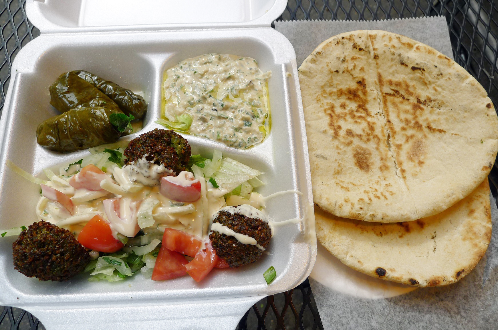 A styrofoam takeout container overflows with dolmas, falafel, and other dishes beside a stack of fluffy pita.
