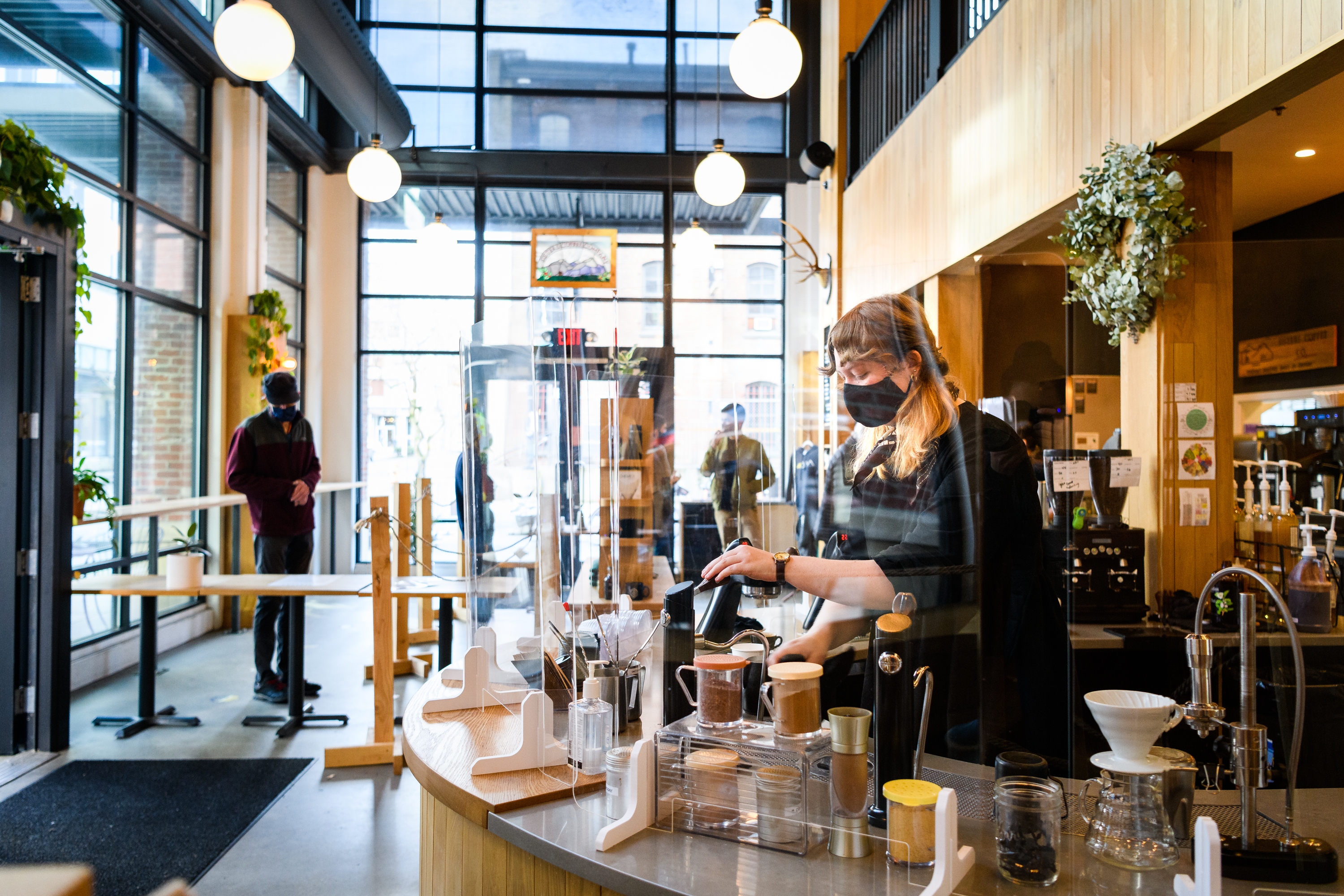 A woman in blonde braids and a black mask makes coffee drinks behind the curved bar at Sisters Coffee, which has high ceilings and globe lights.