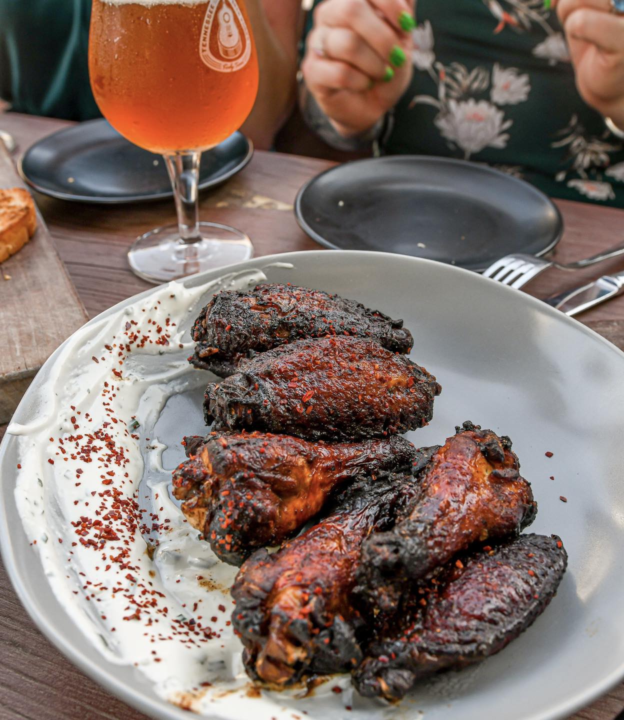 Charred and saucy chicken wings sitting on a white plate with a dusting of cayenne spice. A glass of beer sits behind it. 