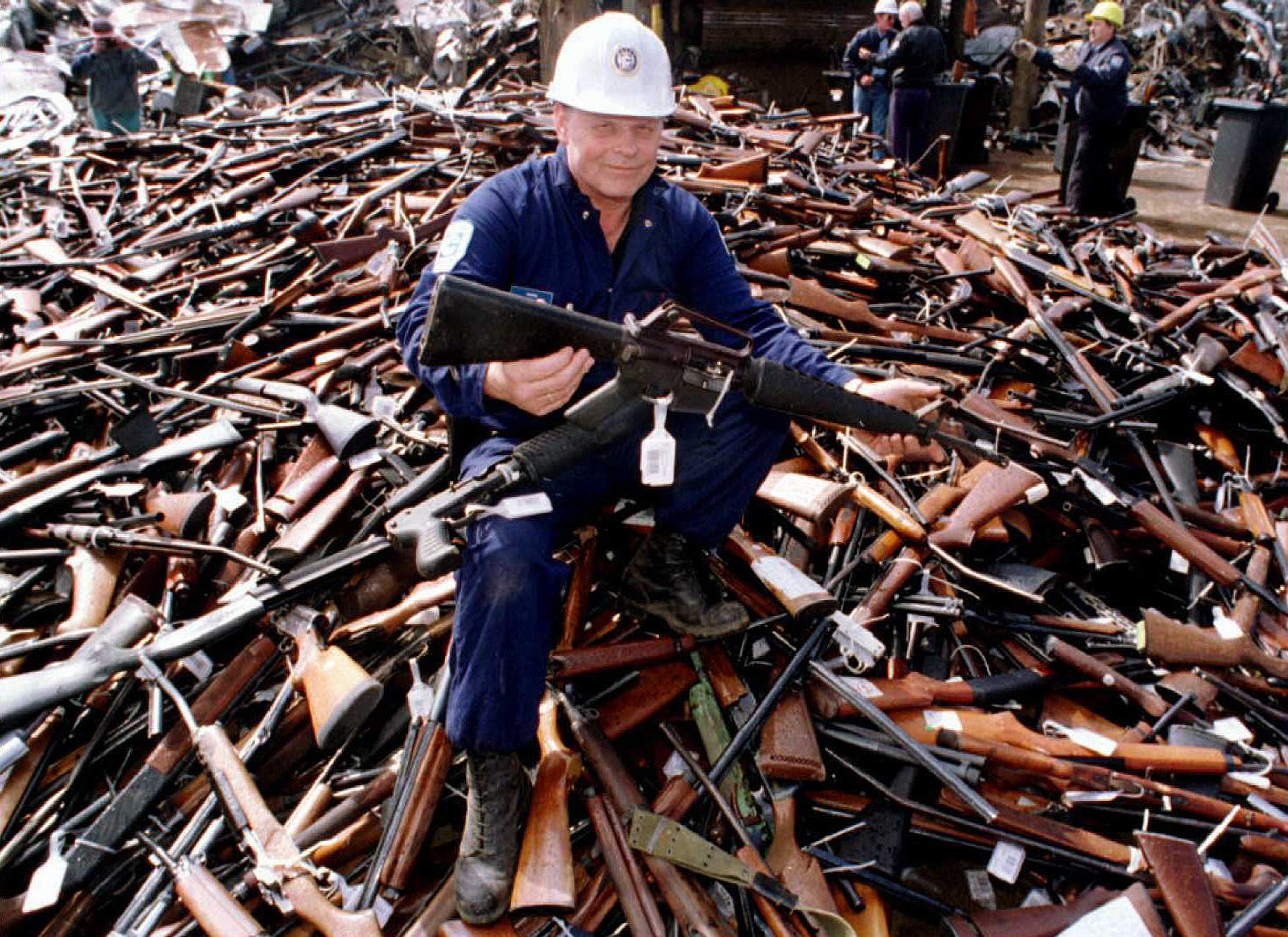 A person wearing a hardhat and holding an automatic rifle sits atop a pile of thousands of confiscated guns.
