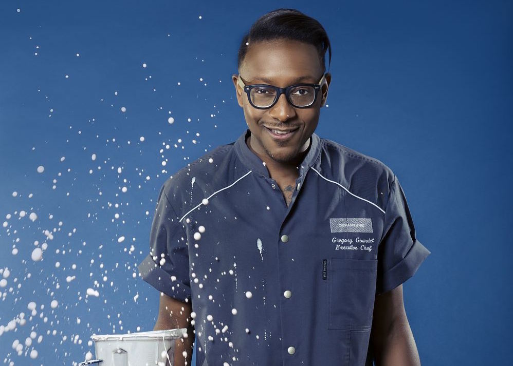 A picture of Gregory Gourdet, the chef at Departure, wearing his uniform as an open blender sprays liquid everywhere