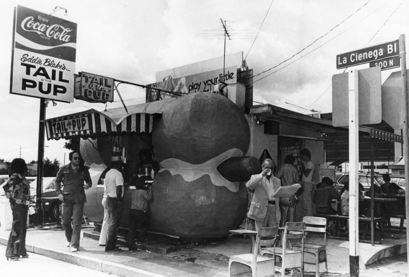 A black and white photo of an old hot dog stand with people waiting to order. The stand is shaped itself like a hot dog.