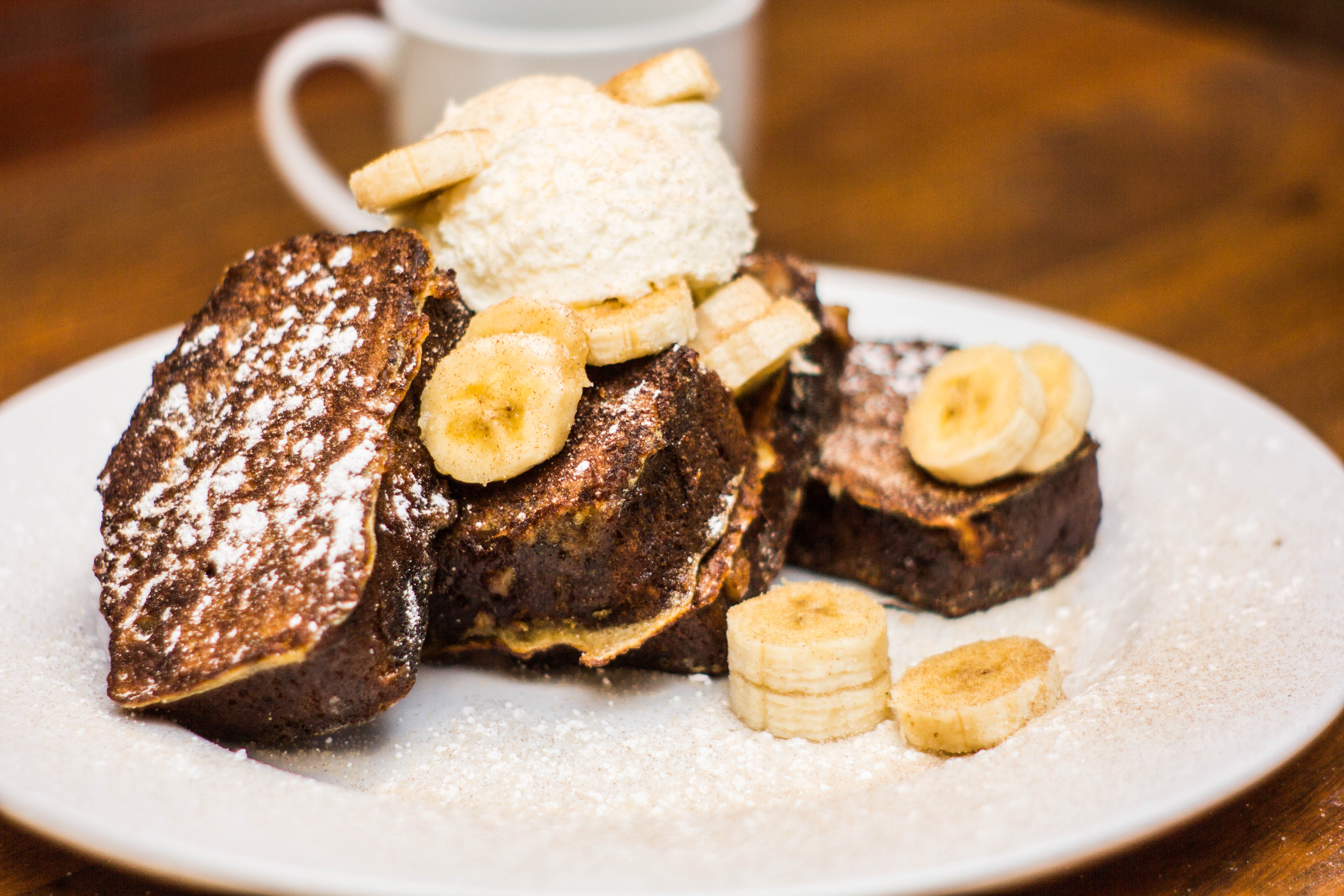 A plate a French toast made with banana bread is embellished with sliced bananas, whipped cream, and powdered sugar. A cup of coffee sits behind it.