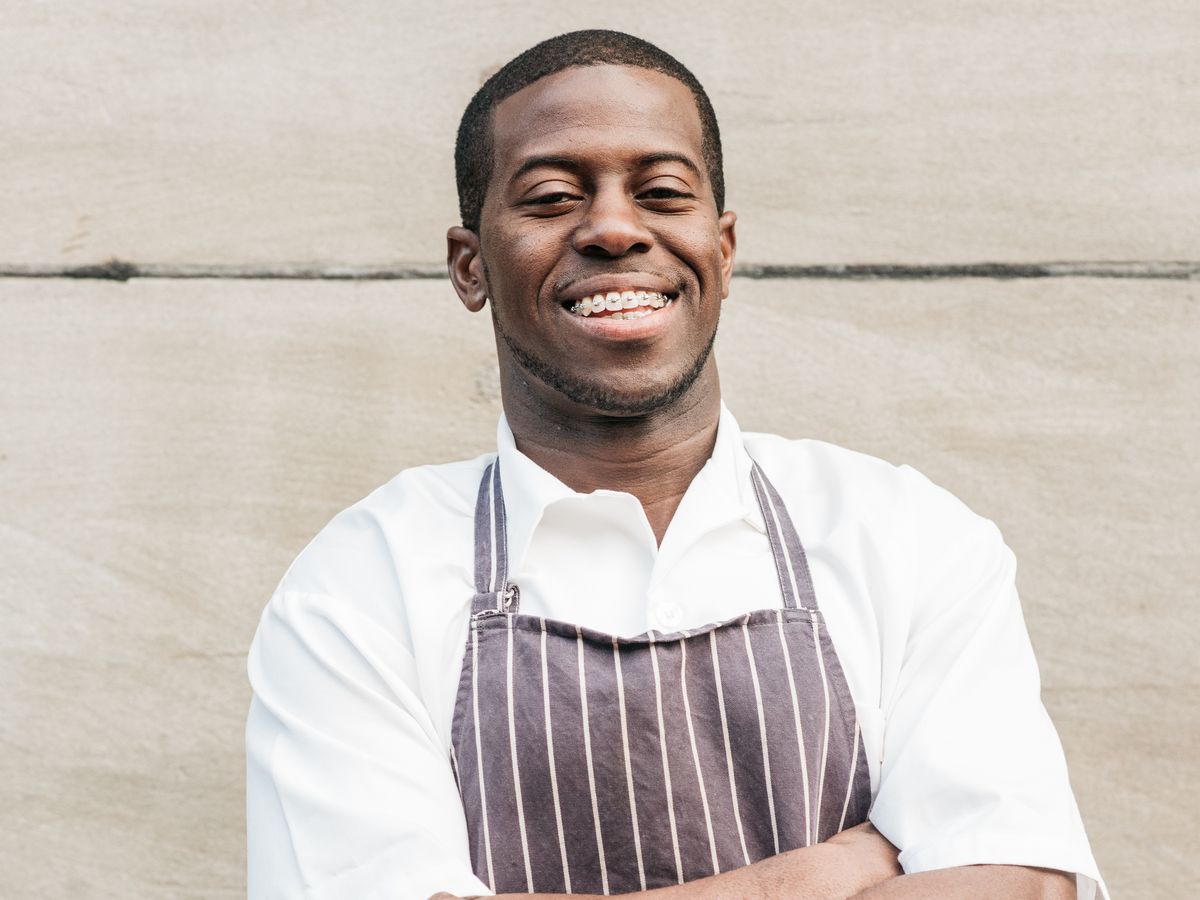 A portrait of chef Edouardo Jordan wearing an apron with his arms folded against a stone wall