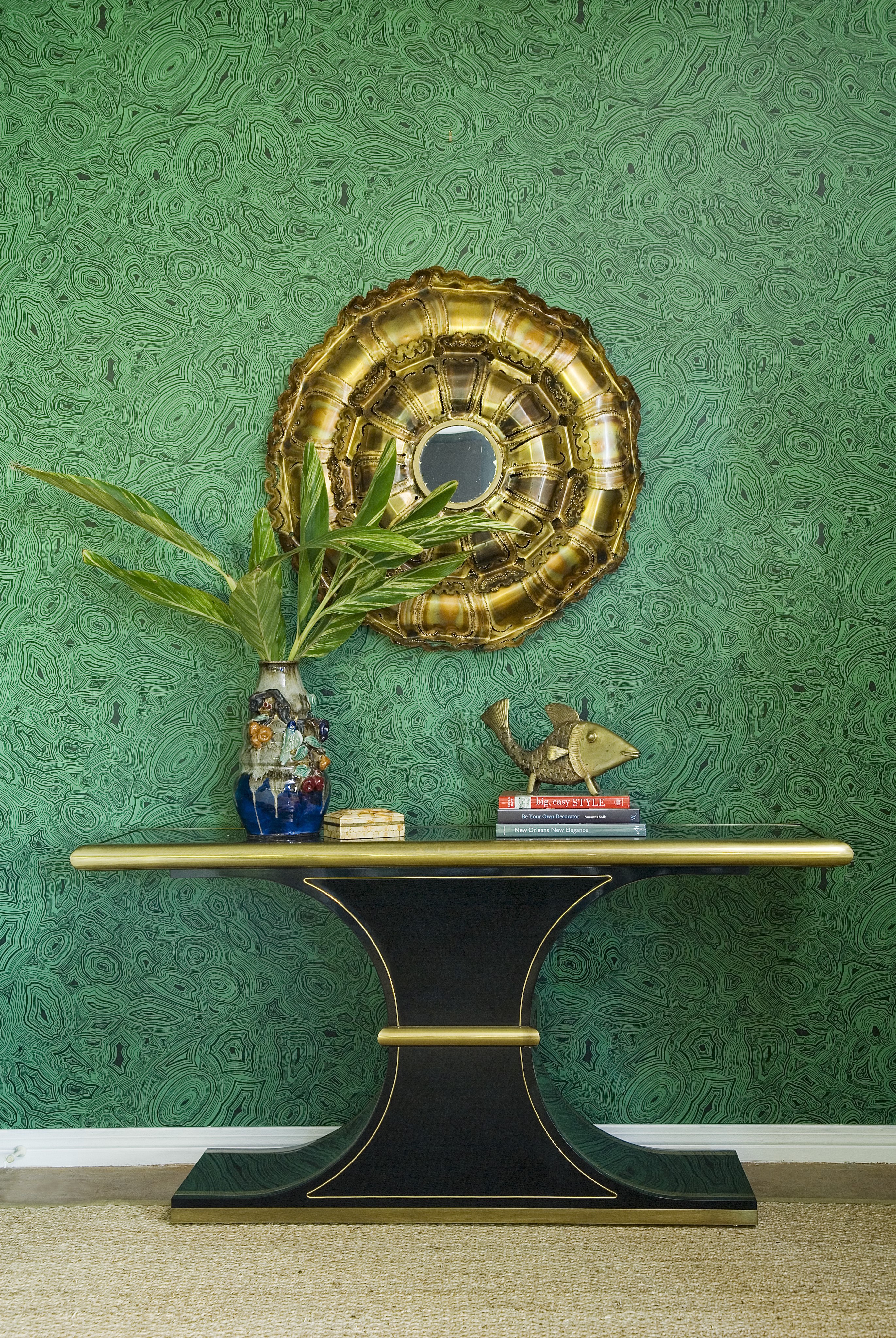 A shelf with a vase, books, and a plant in a vase. The shelf is against a green wall. There is a gold mirror on the wall. 