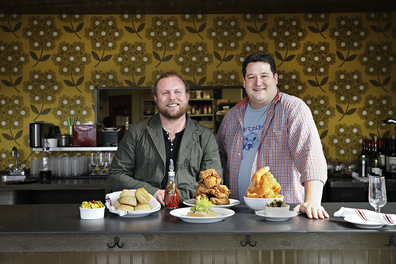 Nick Rancone is on the left, curly blonde hair in a pony tail wearing a suit jacked over a black Henley, Thomas Boemer on the right, with close cropped brown hair, a plaid shirt unbuttoned over a blue t-shirt. In front of them is a spread of Revival dishes including crispy fried chicken.
