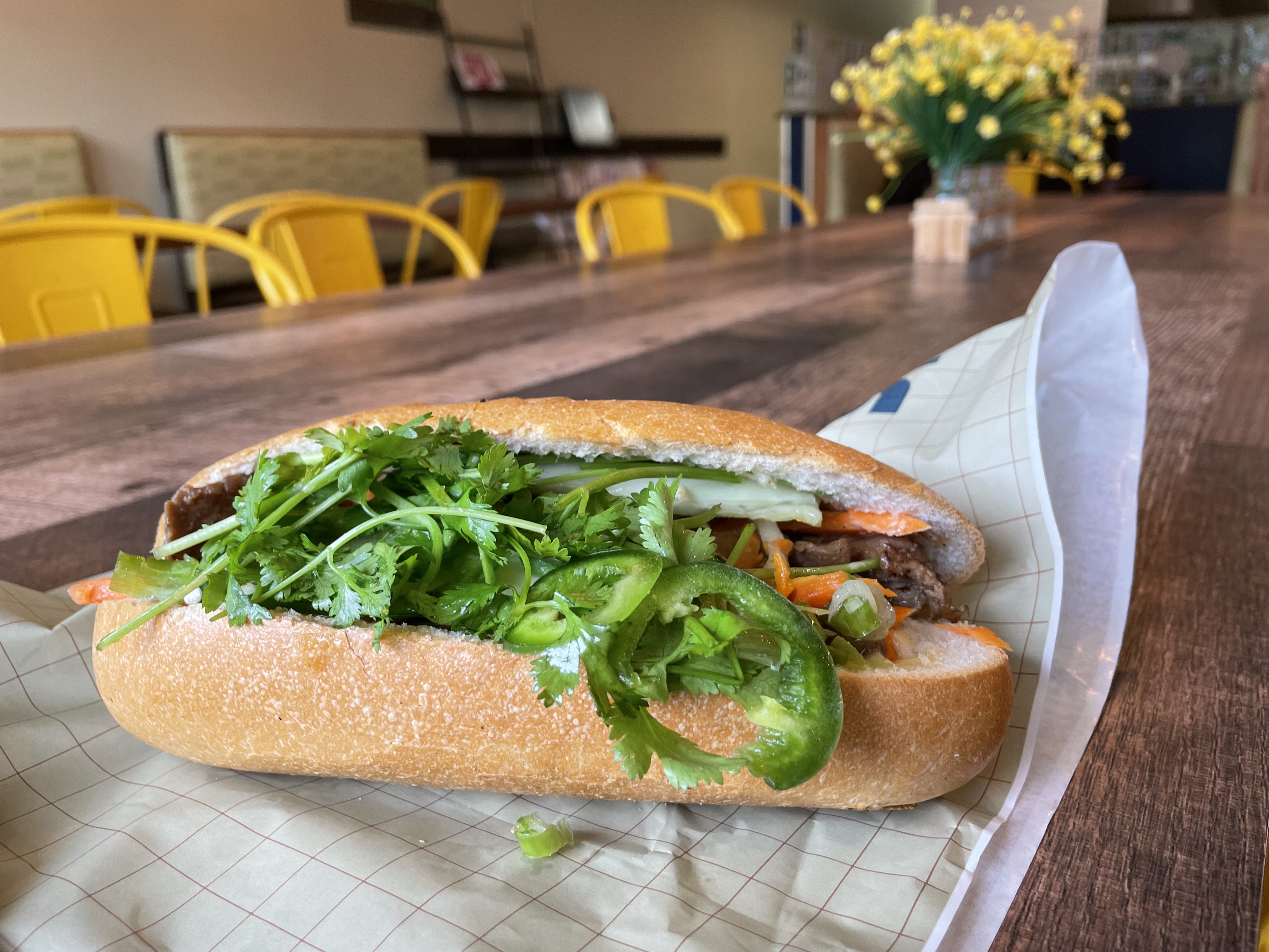 A banh mi sandwich on a wooden table with flowers and yellow chairs in the background. 