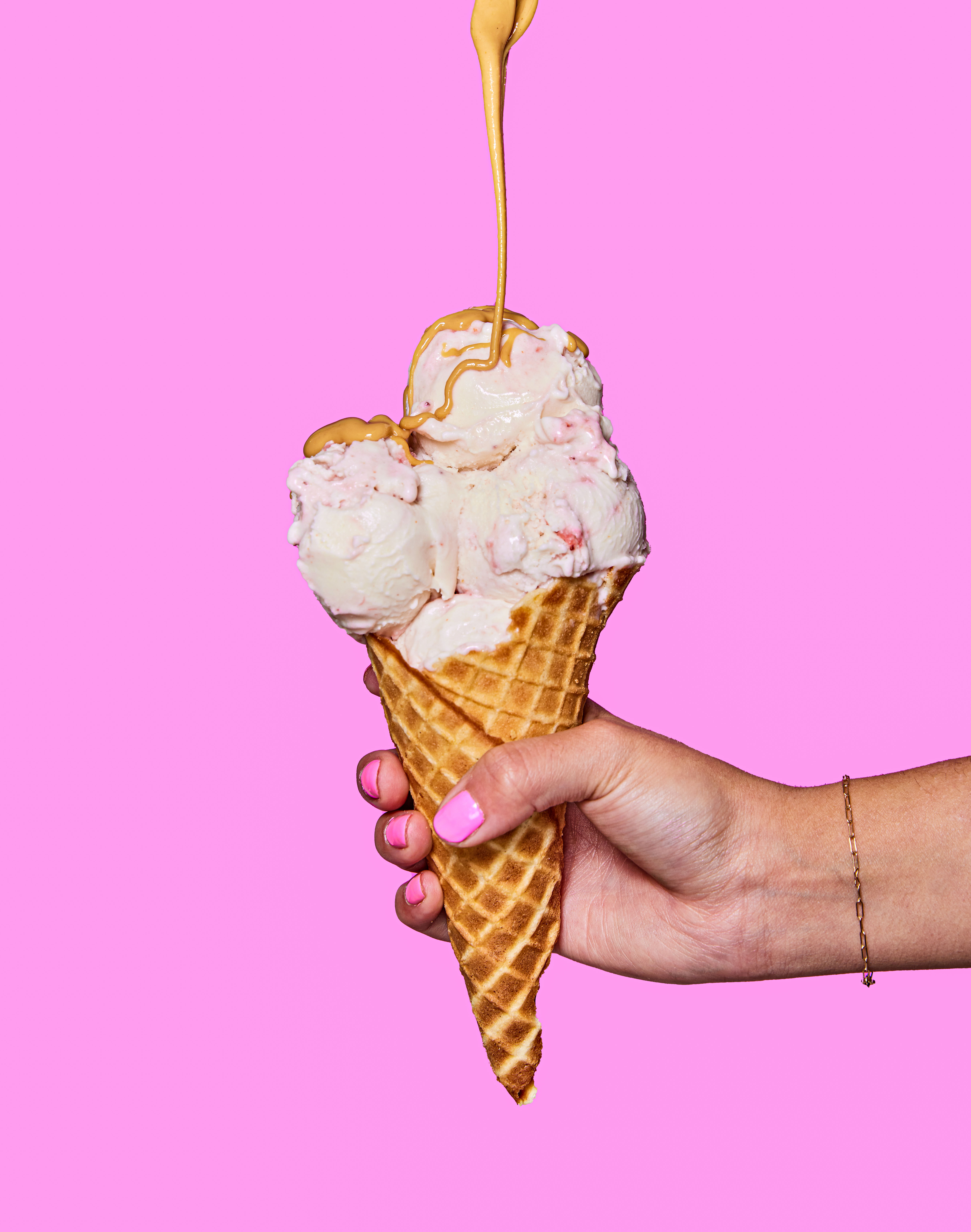 a hand holding a waffle cone topped with three scoops of ice cream with a peanut butter drizzle.
