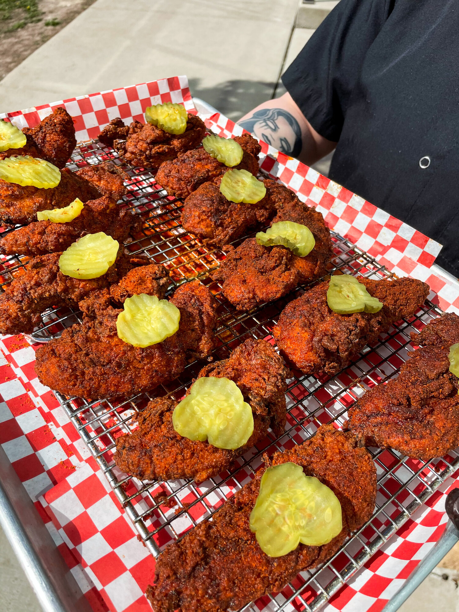 Fried chicken filets on a metal grate above red and white-checkered paper, all topped with a single pickle slice.