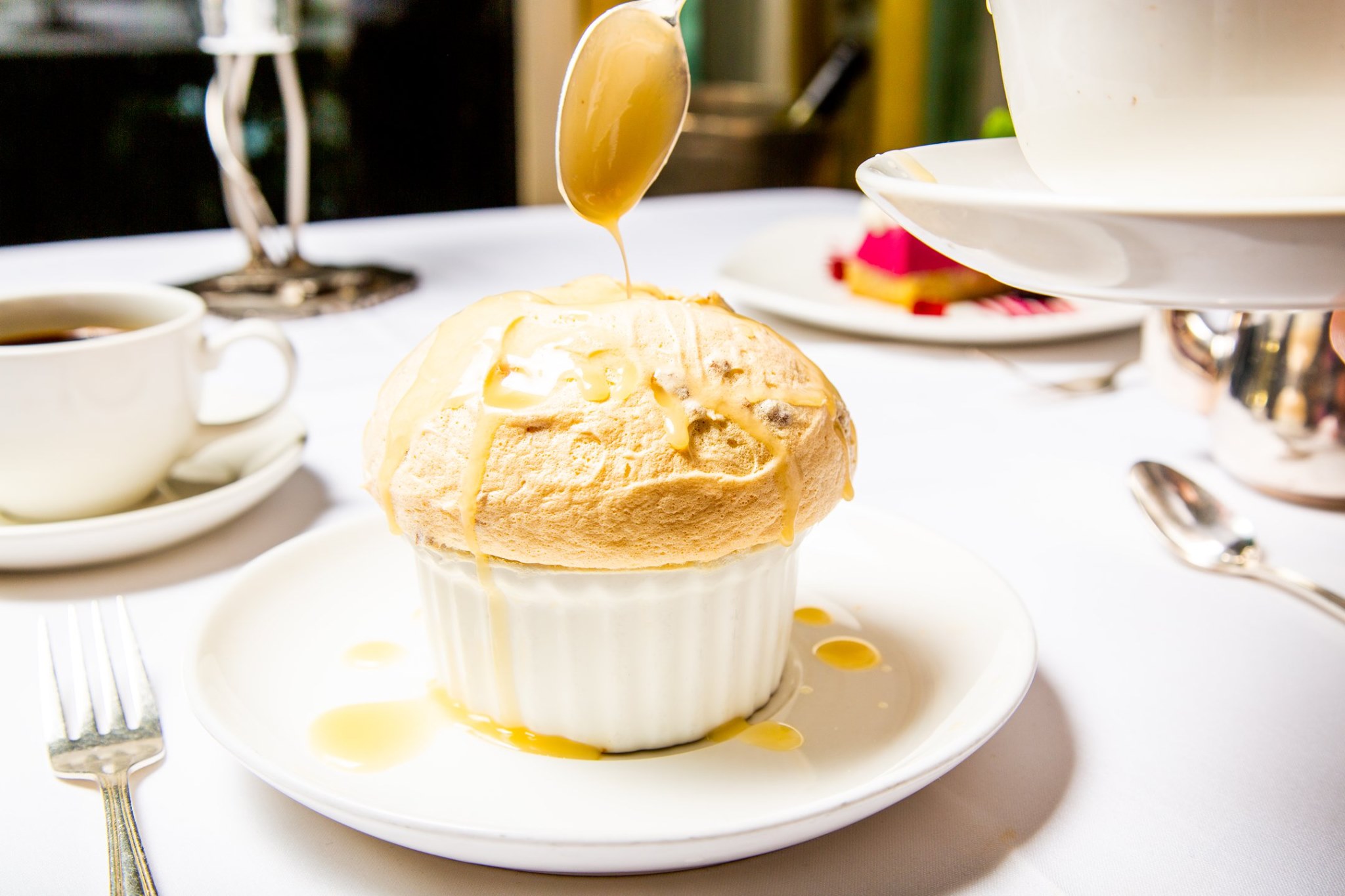 A bread pudding soufflé in a white ramekin on a white plate on a table covered in a white table cloth, with other dishes visible in the background.