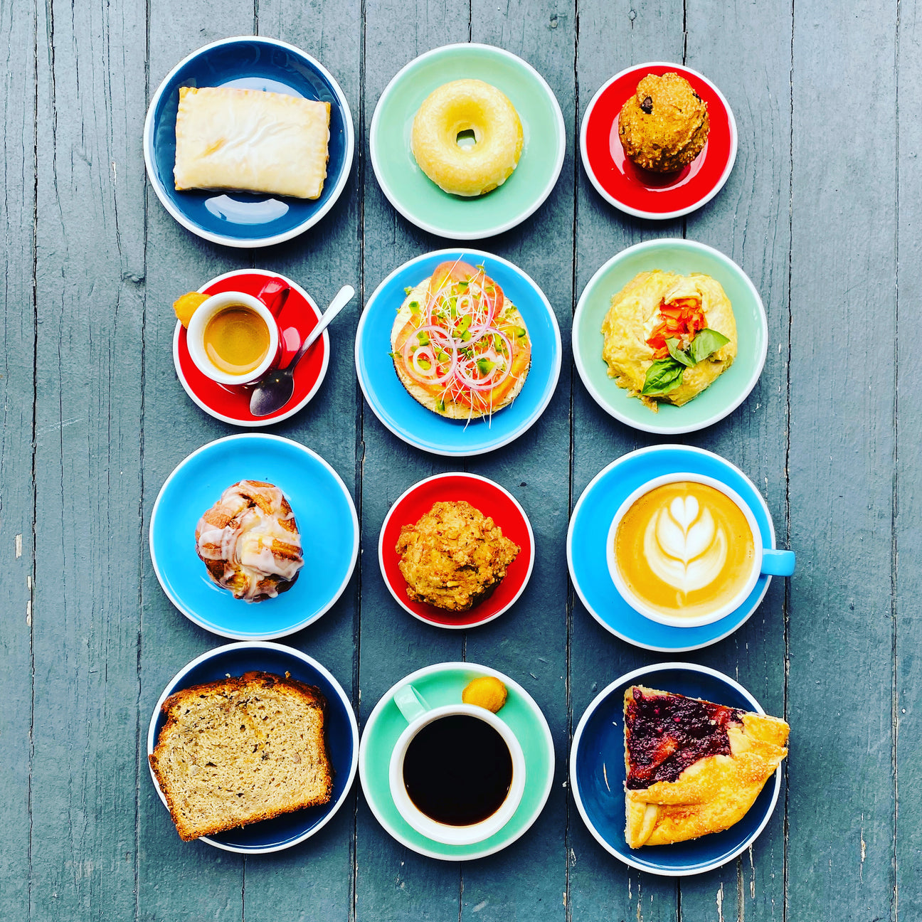 An assortment of pastries and small plates of food next to lattes and espressos in three rows on a grey wooden table, from an aerial view. 