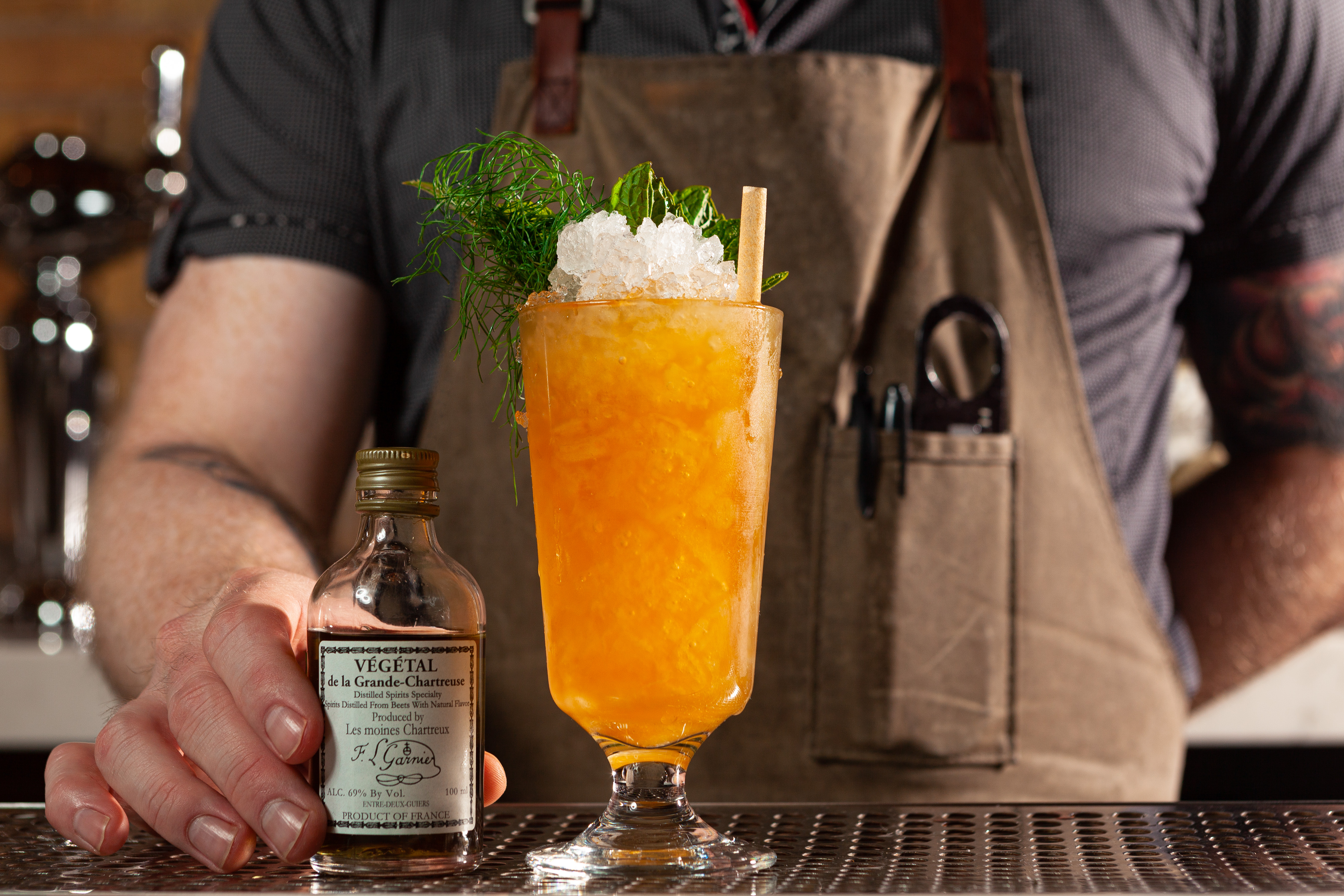 A bartender stands behind an orange cocktail in a tall glass.