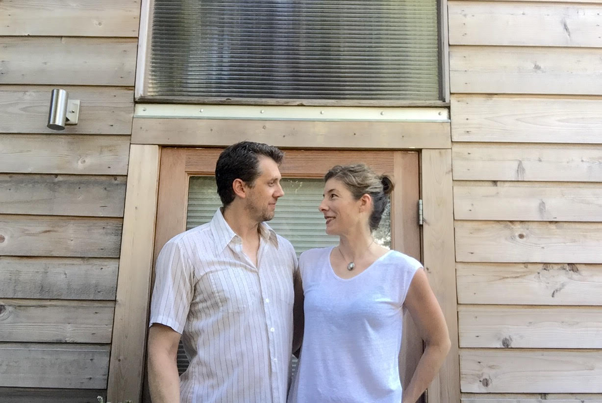 A man on the left and a woman on the right looking at each other lovingly in front of a wood-shingled house 