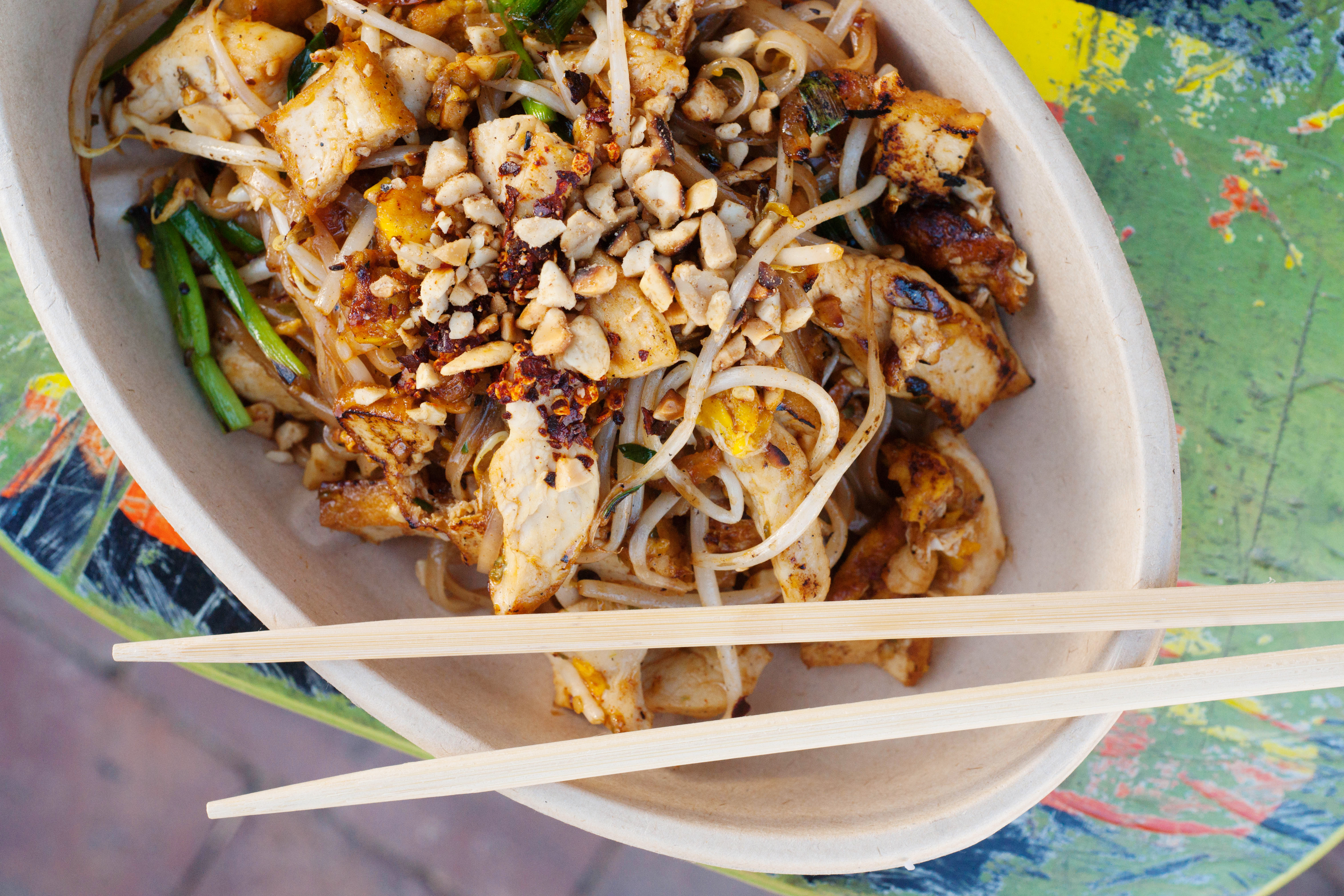 In a paper bowl, a dish of pad thai sits. A pair of chopsticks rest on the rim.